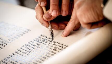 Hands of a scribe holding a quill and touching it to.a Torah scroll.