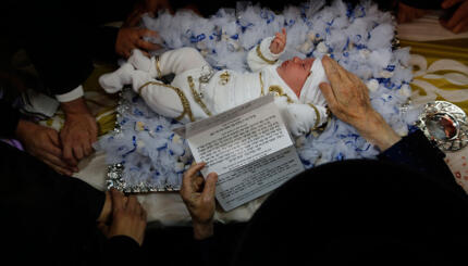 Newborn baby lying on a pillow surrounded by adults, one of whom holds a piece of paper with hebrew writing