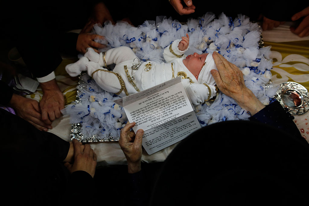 Newborn baby lying on a pillow surrounded by adults, one of whom holds a piece of paper with hebrew writing