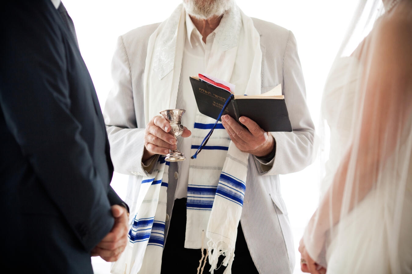 Image of a rabbi's hand holding a kiddush cup between two people getting married.