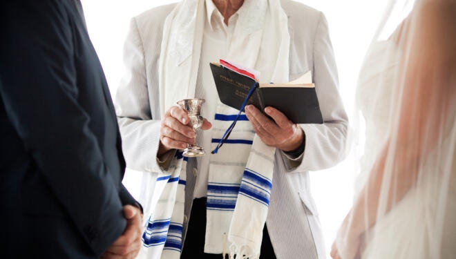 Image of a rabbi's hand holding a kiddush cup between two people getting married.