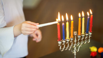 A child uses the shamash to light the Hanukkah candles. The candles are inside a silver menorah.