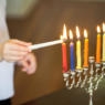 A child uses the shamash to light the Hanukkah candles. The candles are inside a silver menorah.