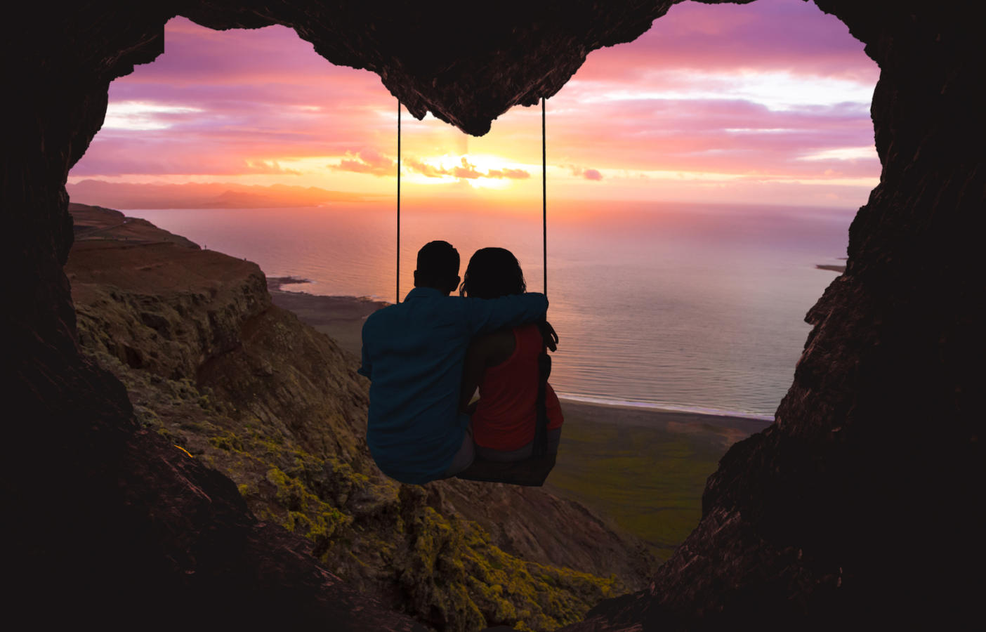 Couple on swing contemplating the sunset over the sea in a romantic view with heart shape.