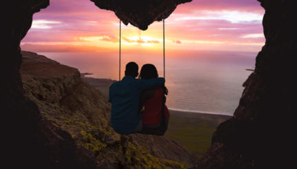 Couple on swing contemplating the sunset over the sea in a romantic view with heart shape.