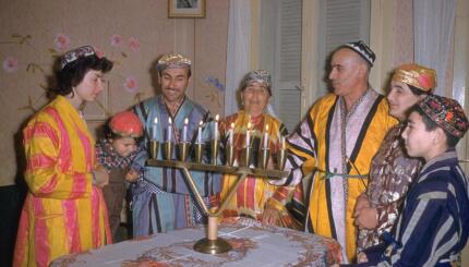 Jews in colorful costume cluster around a Hanukkah menorah.