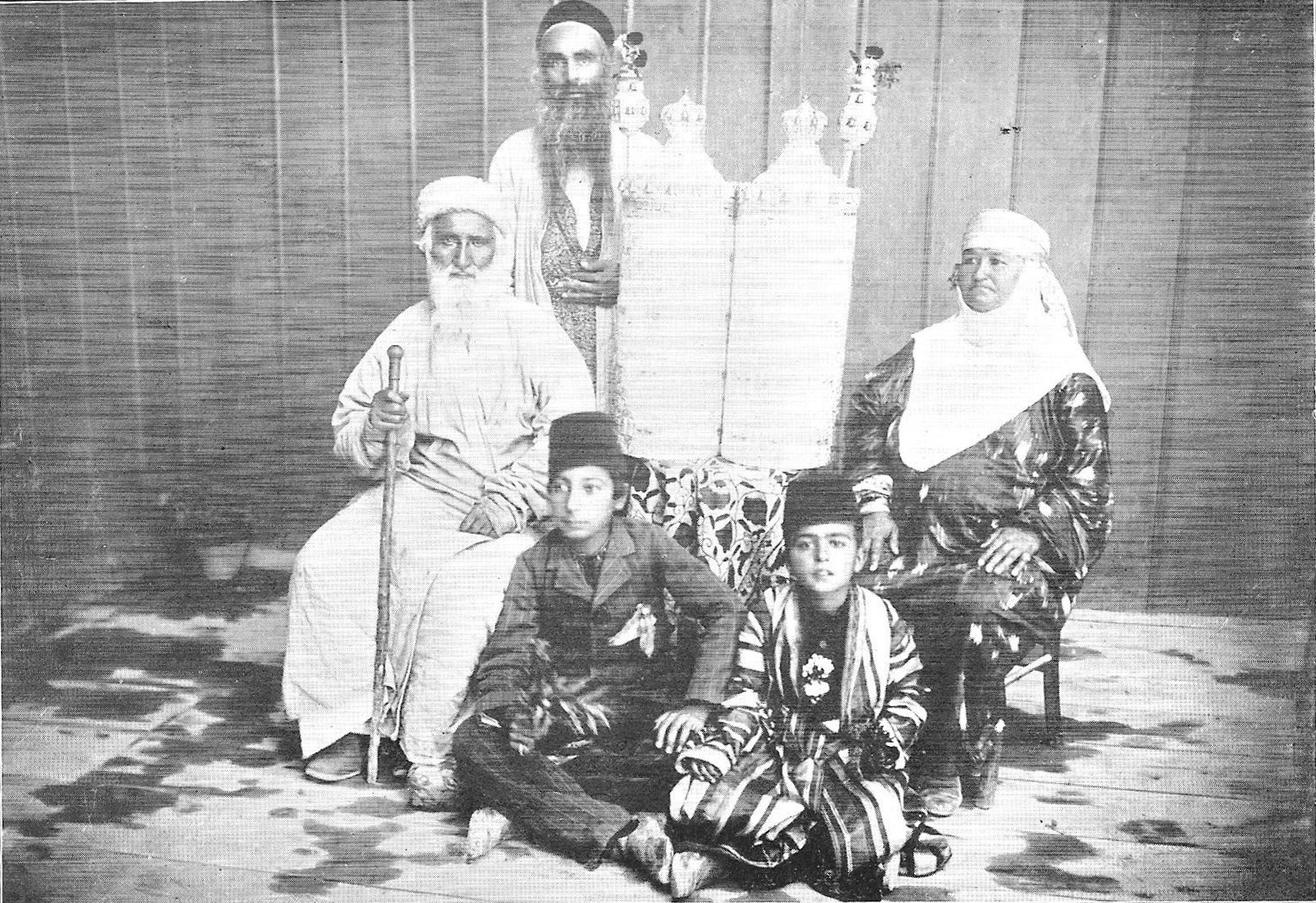 Black and white photo of Jews in traditional Bukharan dress with a Bukharan Torah scroll.