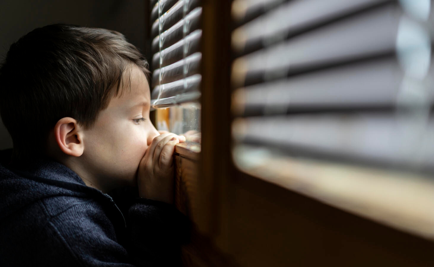 Small sad boy looking through the window during Coronavirus isolation.