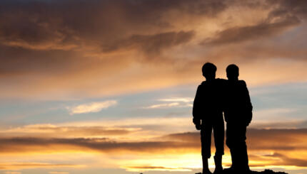 Two happy young boys on a hill at sunset. Back view. Children are elementary aged and have their arms around each other in embrace. Unrecognizable models. Themes in the image include best friends, love, relationships, siblings, brothers, bonding, care, comfort, talking, outside, nature, recreation, leisure, watching the sunset, beauty, and family.