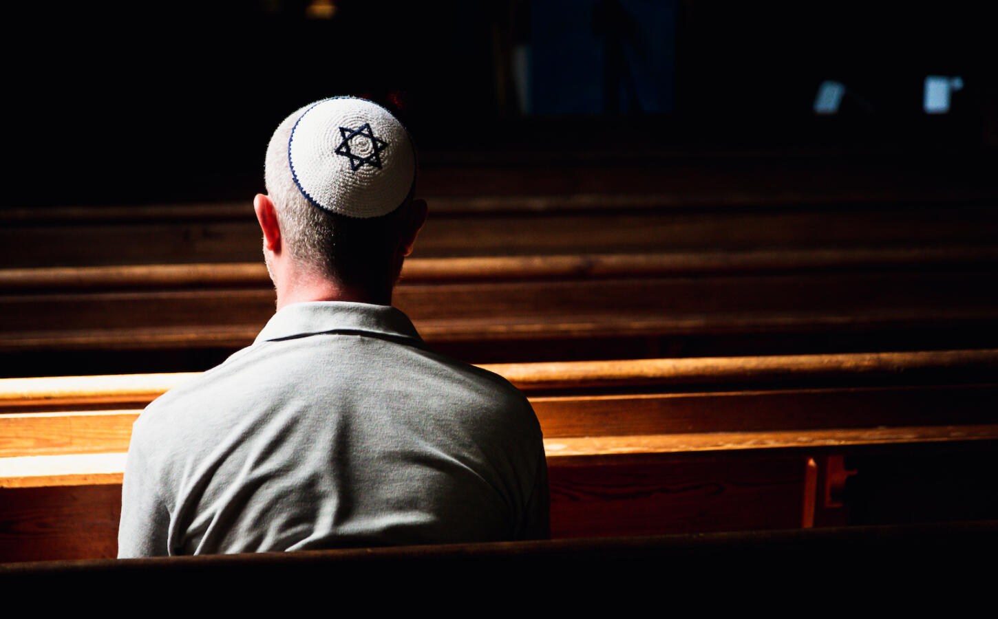 Close up image depicting a young caucasian Jewish adult man in his 30s inside a synagogue. He has his head bowed in prayer and he is wearing the traditional Jewish skull cap - otherwise known as a kippah or yarmulke - on his head. The man has a beard and the background of the synagogue is blurred out of focus. Horizontal color image with copy space.