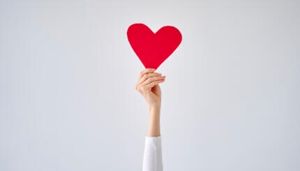 Female's hand holding red heart against white grey background.
