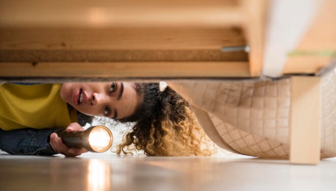 Mixed Race woman looking under bed with flashlight