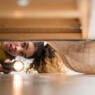 Mixed Race woman looking under bed with flashlight