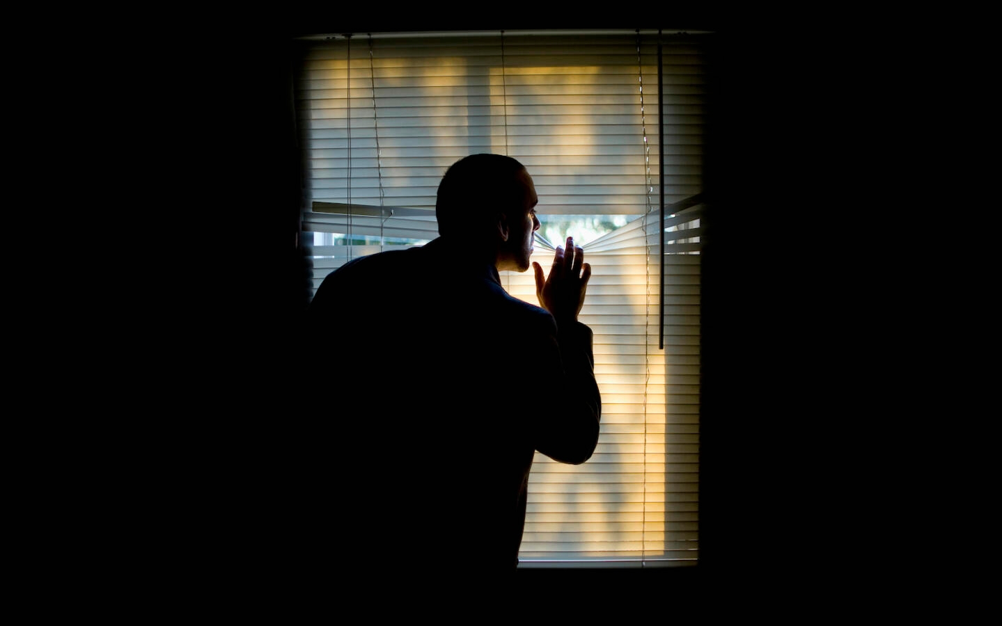 A man peeking outdoors through the blinds of a darkened room