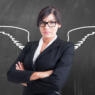 Photo of a woman standing at a blackboard with crossed arms and angel wings drawn behind her.