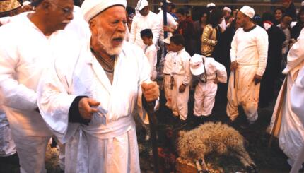 Man in a robe with an intense look on his face next to a slaughtered sheep with a crowd of peple.