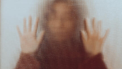 Back lit image of the silhouette of a woman with her hands pressed against a glass window. The silhouette is distorted, and the arms elongated, giving an alien-like quality. The image is sinister and foreboding, with an element of horror. It is as if the 'woman' is trying to escape from behind the glass.
