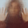 Back lit image of the silhouette of a woman with her hands pressed against a glass window. The silhouette is distorted, and the arms elongated, giving an alien-like quality. The image is sinister and foreboding, with an element of horror. It is as if the 'woman' is trying to escape from behind the glass.